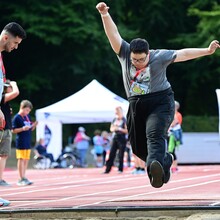 Zwei Männer stehen mit Maßband am Rand einer Sprunggrube und beobachten den Sprung einer männliche Person. Im Hintergrund ein Sportplatz mit weiteren Personen