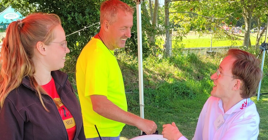 ein Team aus drei Personen an einem kleinen Stand im Schatten auf der Wiese