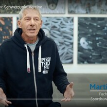 ein Mann in Sportkleidung steht vor einer künstlerisch gestalteten Wand und spricht