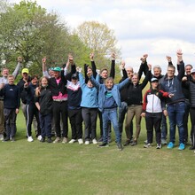 eine große Gruppe von Menschen mit und ohne Behinderungen steht winkend dem grünen Rasen am Rande eines Golfplatzes