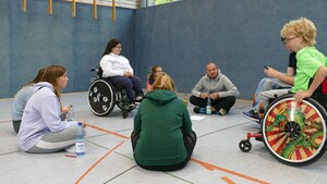 eine Gruppe von Sportlerinnen sitzt zum Teil im Rollstuhl, zum Teil auf dem Boden und diskutiert 