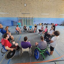 eine Gruppe von Rollstuhlfahrer*innen bildet in einer Turnhalle einen Kreis, drei Personen stehen außerhalb, einer davon fotografiert, die anderen erklären 