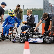 ein Jugendlicher im Rollstuhl sitzt wartend neben dem Kart, das gerade fertig von Monteuren fertig gemacht wird