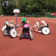 Sechs Kinder in Rollstühlen auf einem Sportplatz um einen aufgestellten Korb mit einem Erwachsenen, der ebenfalls im Rollstuhl sitzt und zwei Basketbälle in den Händen hält