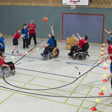 Rollstuhlfahrer*innen und Fußgänger in blauen und roten Trikots spielen unter dem Korb Basketball 
