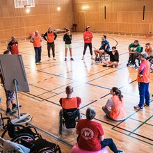 eine Gruppe von Erwachsenen steht in einer Sporthalle im Kreis vor einem Flipchart