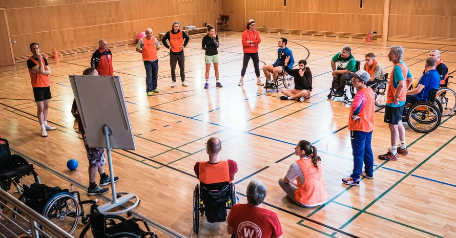 eine Gruppe von Erwachsenen steht in einer Sporthalle im Kreis vor einem Flipchart