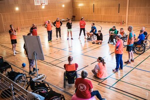 eine Gruppe von Erwachsenen steht in einer Sporthalle im Kreis vor einem Flipchart