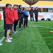 ein Mann (Taime Kuttig) steht auf einem Fußballfeld vor einer Gruppe von Männern in Sportkleidung