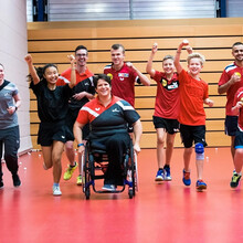 Eine Gruppe von Sportler*innen unterschiedlichen Alters läuft mit viel Spaß in einer Sporthalle, im Vordergrund eine Rollstuhlfahrerin