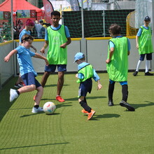 sechs Kinder und Jugendliche, vier davon in hellgrünen Leibchen, spielen Fußball auf einem von Banden begrenzten Spielfeld