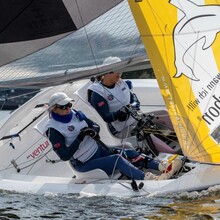 zwei Personen mit norwegischer Flagge auf dem Trikot in einem schiefliegenden Segelboot mit gelbem Segel, auf dem ein Delfin abgebildet ist. 