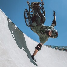 ein Rollstuhlfahrer mit Helm und Ellenbogenschützern fliegt durch eine Halfpipe, der Rollstuhl und die Beine befinden sich gerade oberhalb von Kopf und Schultern, eine Hand berührt den Beton