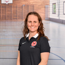 eine junge Frau im schwarzen Polo-Hemd mit Vereinsenblem in einer Sporthalle