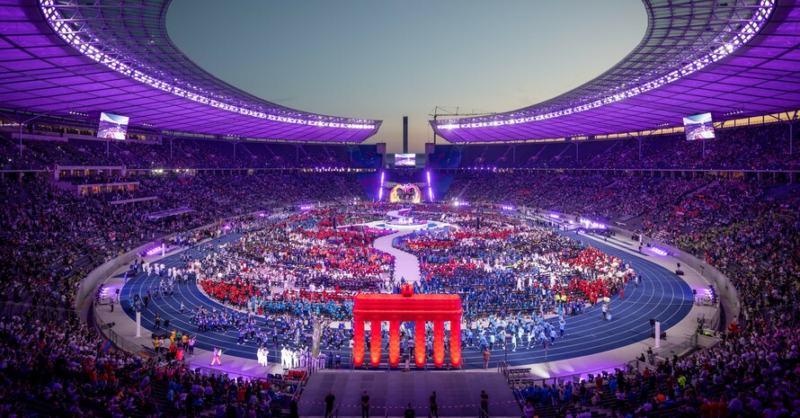 Blick von oben in ein volles Stadion in lila Licht, im Vordergrund ein Nachbau des Brandenburger Tors in rot