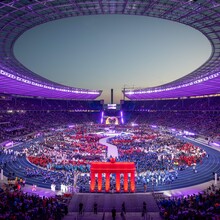 Blick von oben in ein volles Stadion in lila Licht, im Vordergrund ein Nachbau des Brandenburger Tors in rot