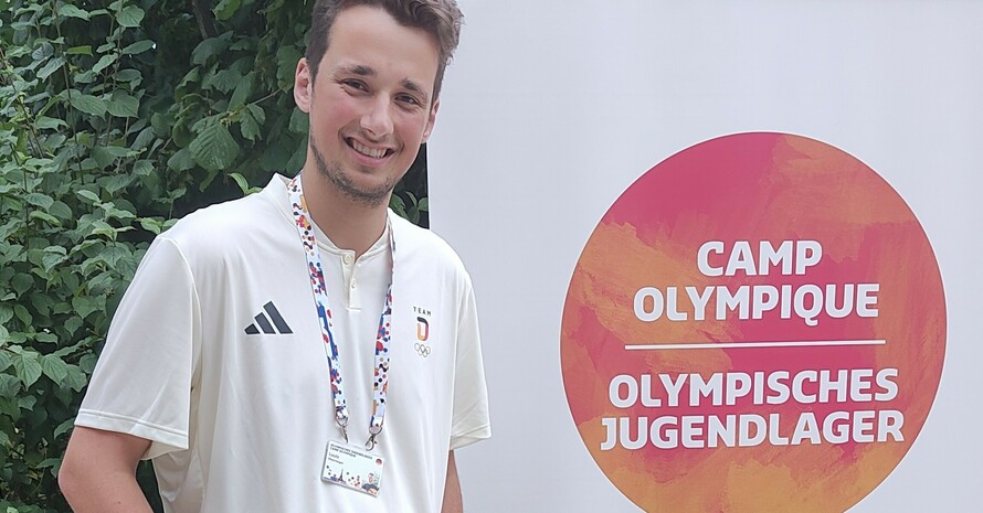 Ein junger Mann mit Akkreditierung an einem blauen Band um den Hals steht vor einem Banner mit der Aufschrift Olympisches Jugendlager in deutscher und französischer Sprache