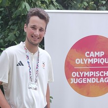 Ein junger Mann mit Akkreditierung an einem blauen Band um den Hals steht vor einem Banner mit der Aufschrift Olympisches Jugendlager in deutscher und französischer Sprache