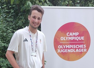 Ein junger Mann mit Akkreditierung an einem blauen Band um den Hals steht vor einem Banner mit der Aufschrift Olympisches Jugendlager in deutscher und französischer Sprache
